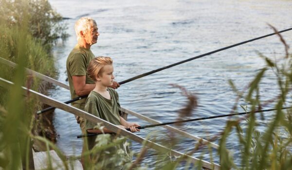 european grey haired mature father with son outdoors fishing by lake river standing near water with fishing rods hands dress casually enjoying hobby nature