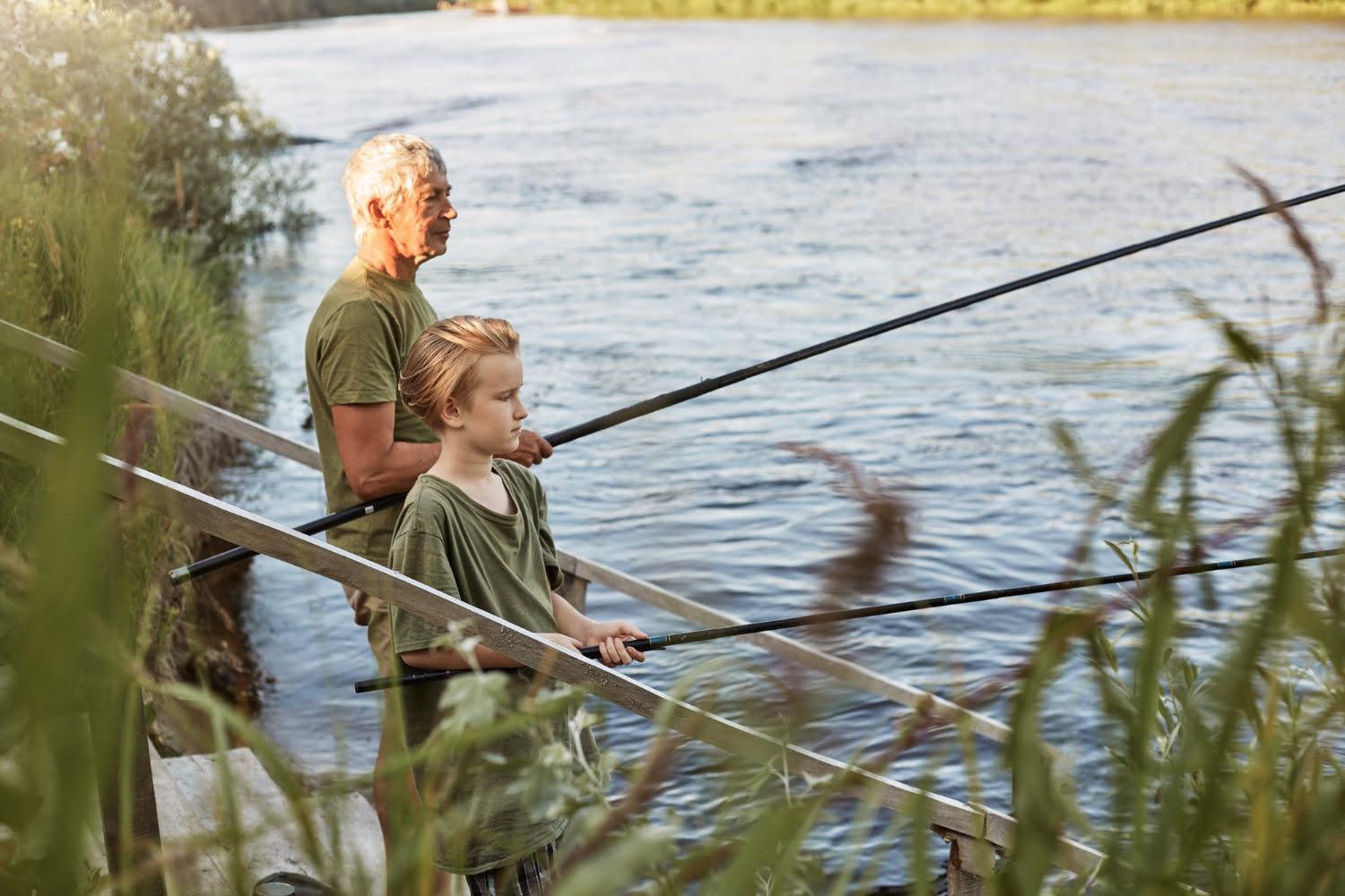 european grey haired mature father with son outdoors fishing by lake river standing near water with fishing rods hands dress casually enjoying hobby nature